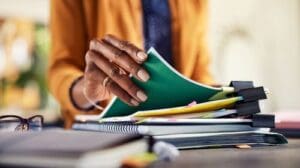 A person sorts through a stack of colorful folders and papers on a desk, with notebooks and paper clips visible in the foreground.
