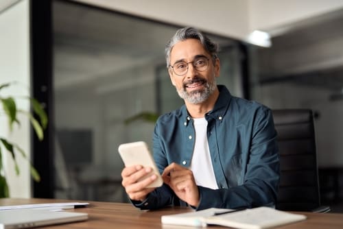 A man with glasses and a beard sits at a desk, smiling while holding a smartphone, with a notebook and laptop in front of him.