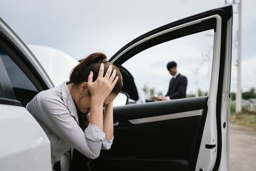 A woman sits in a car with her head in her hands, looking distressed, while a man stands outside in the background near another car.
