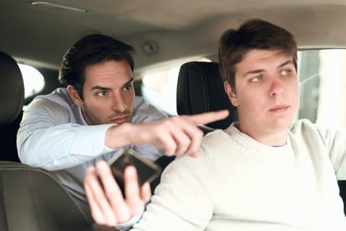 Two men are sitting in a car. The man in the back seat is gesturing and appears to be upset, while the driver looks away with a neutral expression.