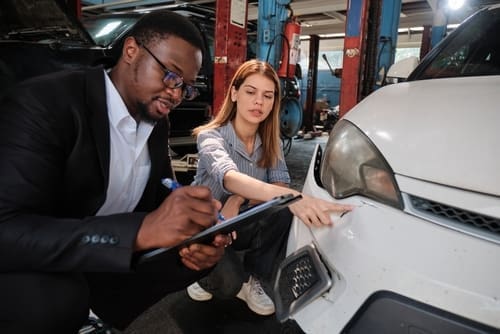 A man in a suit writes on a clipboard while a woman points at damage on the front bumper of a white car in an auto repair shop.