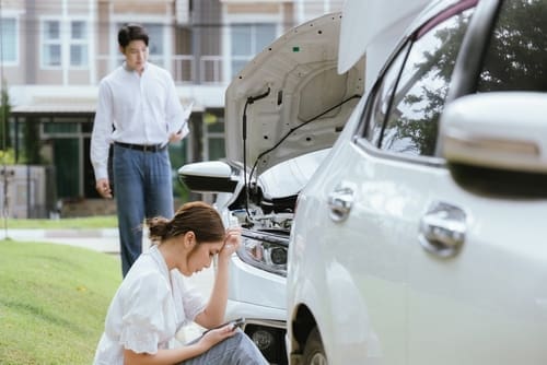 A woman sits on the grass looking at her phone near a car with the hood open, while a man stands in the background holding papers.