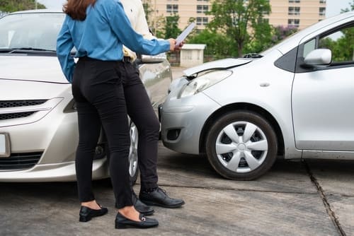 Two people stand near two cars that appear to have been in a minor accident, discussing or reviewing something on a tablet.