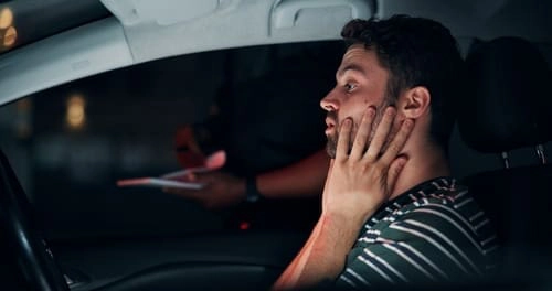A man sits in a car at night, looking concerned with his hands on his face, while a person outside holds a notepad or paper.