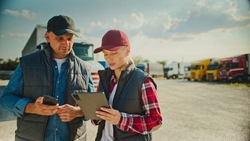 Two people wearing caps and vests stand outdoors near trucks, looking at a tablet and a phone, possibly discussing logistics or transportation details.