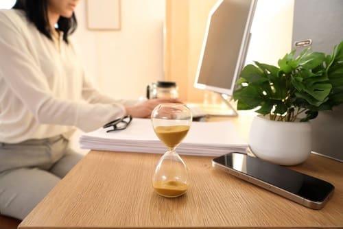 A person works at a desk with a computer, stack of papers, and coffee mug; an hourglass, smartphone, and potted plant are in the foreground.