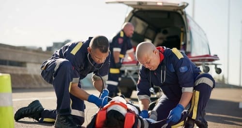 Two paramedics provide emergency medical care to a person lying on the road near an open ambulance, with another responder in the background.