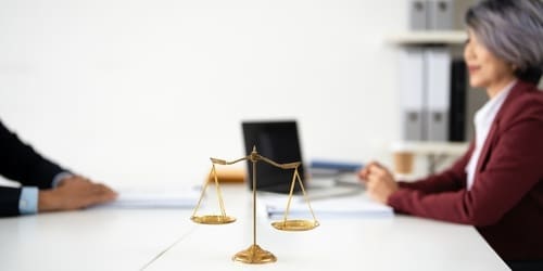 A brass balance scale sits on a table between two people in business attire, with office folders and a laptop in the background.