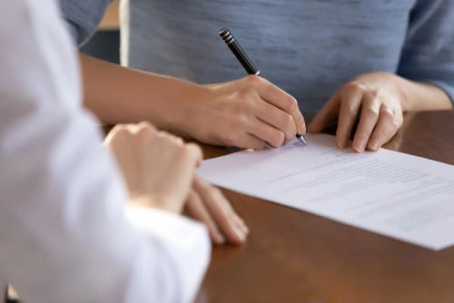Two people sitting at a table, with one person holding a pen and signing a document while the other person observes.