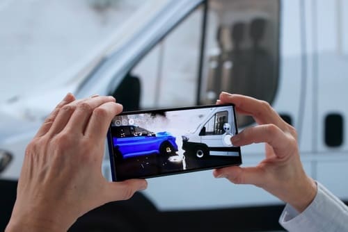 A person holds a smartphone taking a photo of a car accident involving a blue car and a white van.