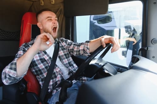 A man wearing a seatbelt is sitting in the driver’s seat of a vehicle, yawning with his eyes partly closed and one hand on the steering wheel.