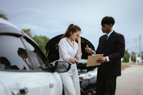 A woman stands by a damaged car with the hood open, looking distressed. A man in a suit holds a clipboard and speaks to her on the roadside.
