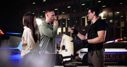 Police officer administering blood test to seated man inside patrol vehicle