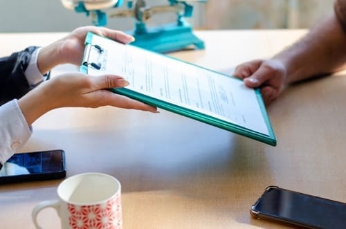 Two people exchange a clipboard with documents across a table; smartphones, a cup, and a scale are also visible.