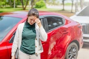 A woman stands next to a red car with a damaged side, holding her head with a distressed expression.