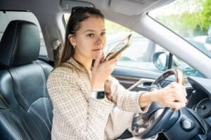 Woman sitting in the driver’s seat of a car, holding the steering wheel with one hand and speaking into her smartphone with the other.