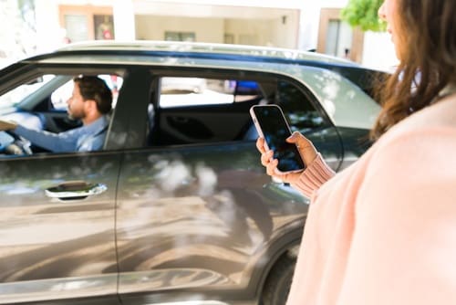 A woman holding a smartphone stands next to a parked car while a man is seated in the driver’s seat.