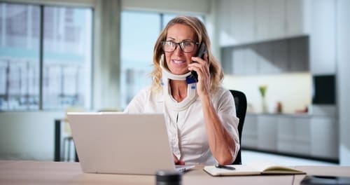 A woman wearing a neck brace sits at a desk, talking on the phone and using a laptop, with a notebook and pen nearby in a bright, modern home office.