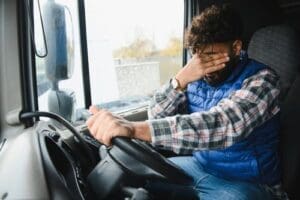 A man sitting in a truck driver's seat rests his hand on the steering wheel and covers his face with his other hand, appearing tired or stressed.