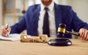 A person in a suit uses a gavel next to wooden toy cars on a desk, suggesting a legal decision related to automobiles.