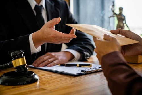Two people in business suits sit at a desk with a gavel, clipboard, and pen, as one hands an envelope to the other.