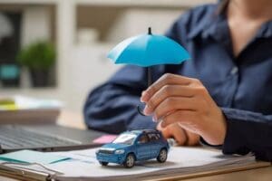 A person holds a small blue umbrella over a toy car on a desk, symbolizing car insurance or vehicle protection.