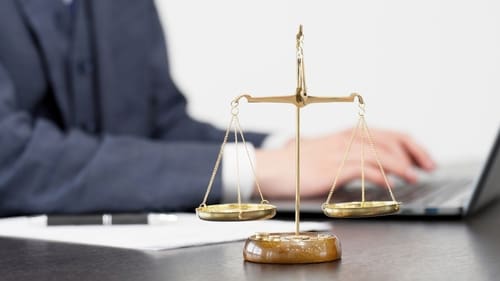 A brass balance scale sits on a desk in front of a person using a laptop, symbolizing justice and legal work.