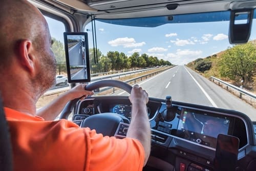 A person drives a truck on a clear highway, with navigation screens and a digital side mirror visible inside the cab.
