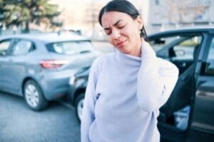 Woman standing outside with a pained expression, holding her neck after a car accident with two vehicles visible in the background.