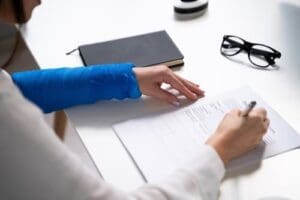 Charlotte Hit and Run Accident Lawyer - Payne Law Firm Person with a blue arm cast is filling out a form at a desk with a notebook and eyeglasses nearby.