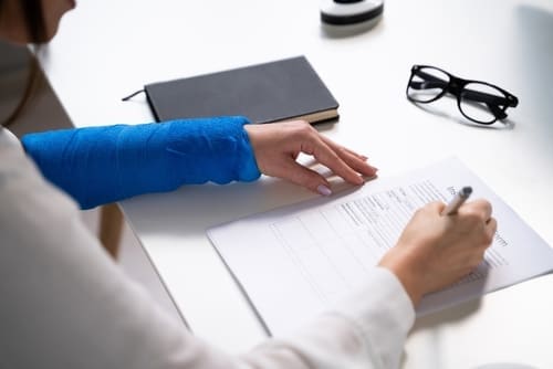 Person with a blue arm cast is filling out a form at a desk with a notebook and eyeglasses nearby.