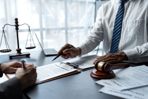 Two people in business attire review and sign legal documents at a desk with a gavel, scales of justice, and papers, in an office setting.