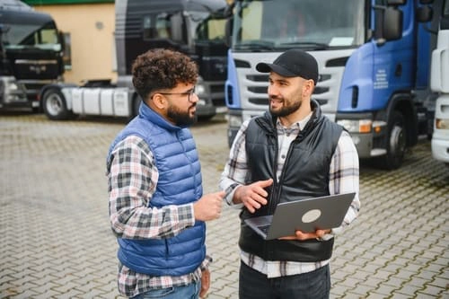 Two men standing outdoors in front of parked trucks, having a discussion; one holds a laptop and gestures while the other listens.