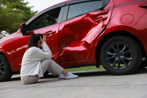 distressed woman sitting on ground beside severely dented vehicle after collision