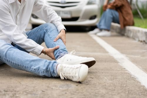 A person sits on the road holding their ankle, appearing injured, with a car and another person in the background.