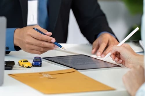 Two people review a digital contract on a tablet at a desk with toy cars, a pen, keys, and a yellow envelope.