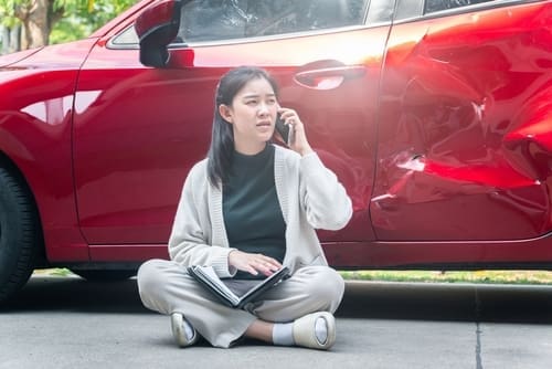 woman sitting on ground calling for help beside dented damaged car