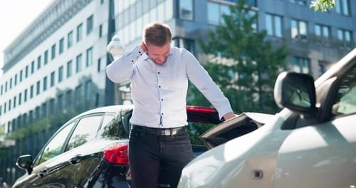 A man stands between two cars on a street, holding his neck and looking at the damage after a car accident.