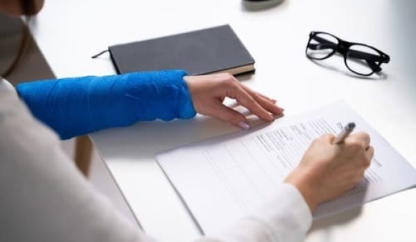Person with a blue arm cast is filling out a form at a desk with a notebook and eyeglasses nearby.