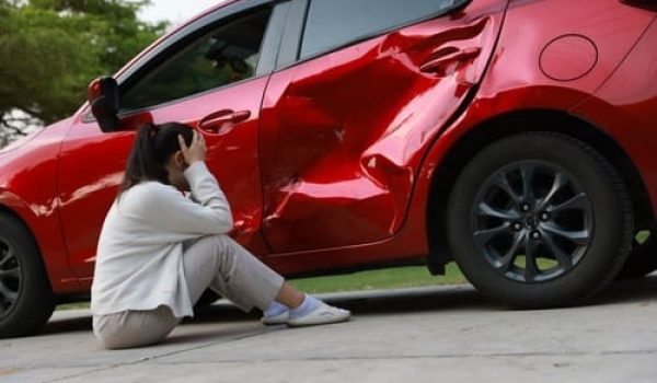 distressed woman sitting on ground beside severely dented vehicle after collision