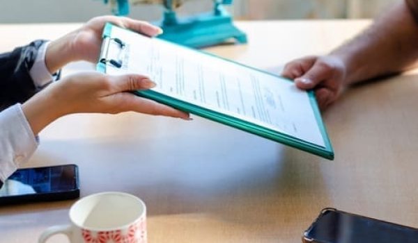 Two people exchange a clipboard with documents across a table; smartphones, a cup, and a scale are also visible.