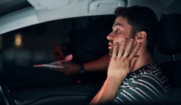 A man sits in the driver’s seat of a car at night, looking concerned, while a person outside the car holds a notepad.