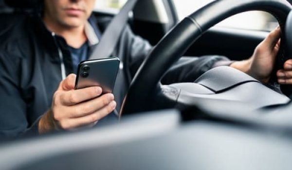 A person sits in the driver's seat of a car, looking at a smartphone and holding the steering wheel with one hand.