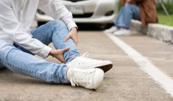 A person sits on the road holding their ankle, appearing injured, with a car and another person in the background.