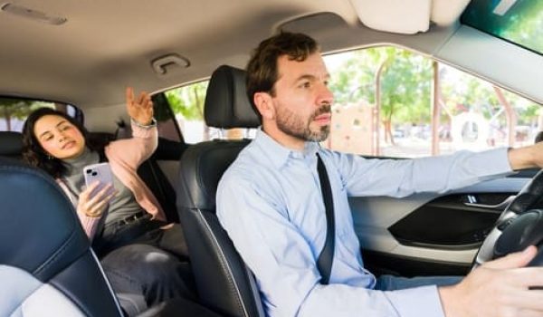 A man drives a car while a woman sits in the back seat, holding a phone and gesturing with her hand.