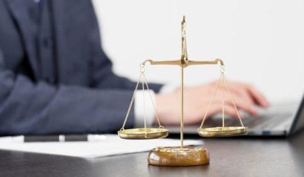 A brass balance scale sits on a desk in front of a person using a laptop, symbolizing justice and legal work.